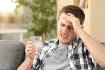 Man suffering head ache with a hand on forehead and holding a glass with a painkiller sitting on a couch at home.